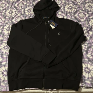 Svart hoodie från Polo Ralph Lauren - Svart hoodie från Polo Ralph Lauren med dragkedja framtill, huva med snörning och klassisk broderad logga på bröstet. Tröjan har kängurufickor och är tillverkad i mjuk bomullsmix. Perfekt för en chill och stilren look.