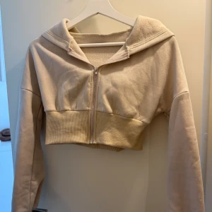 Beige croppad hoodie från SHEIN - Beige croppad hoodie från SHEIN i mjuk polyester. Tröjan har huva, lång ärm och dragkedja framtill. Ribbad bred mudd nedtill ger en snygg passform. Perfekt för en chill och trendig look.
