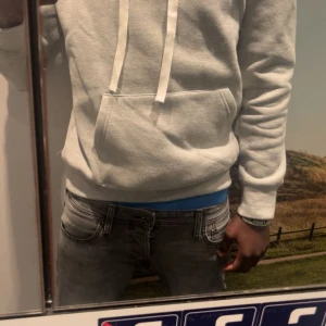 Grå hoodie från Ralph Lauren - Säljer en stilren grå hoodie från Ralph Lauren med klassisk blå logga på bröstet. Tröjan har huva med vita snören och en stor magficka. Perfekt för dig som gillar en enkel och clean look.