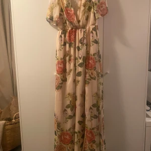 Blommig långklänning från NLY Eve - Romantisk långklänning från NLY Eve i ljusbeige med stora blommor i rosa, grönt och gult. Klänningen har v-ringning, korta ärmar och markerad midja. Tunt, flowy material som ger en drömmig känsla. Perfekt för sommarens alla tillfällen!