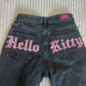 Hello Kitty x Bershka mörkgrå jeans - Unika mörkgrå jeans från Bershka x Hello Kitty med rosa tryck på bakfickorna och rosa strasstenar i flames-mönster vid bensluten. Några har lossnat men det tillkommer extra strass som man kan limma på. Byxorna har midja och rak passform. (Är 1,73 för referens!) jätte fräcka byxor passa på💗