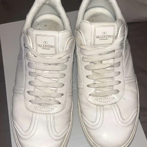 Vita Valentino Garavani sneakers - Snygga helvita sneakers från Valentino Garavani i skinn med klassisk snörning och diskret logga på plösen. Yttersulan har ett unikt camouflagemönster i grönt och svart. Perfekta för dig som gillar stilrena och exklusiva skor med en twist.