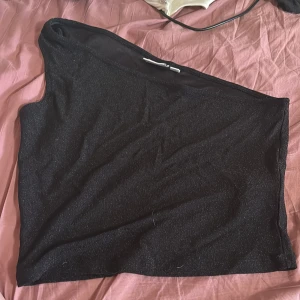 Svart glittrig oneshoulder topp från Vila - Svart glittrig oneshoulder topp från Vila i storlek L. Toppen har en snygg asymmetrisk design med en axel och är perfekt för dig som vill sticka ut. Materialet är mjukt och stretchigt med en diskret glittrig finish som ger extra edge.