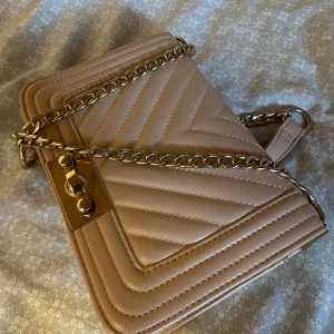 Beige quiltad axelväska med kedja - Snygg beige axelväska med quiltad design och guldfärgad kedjerem. Väskan har ett elegant spänne framtill och stilrena sömmar i mönster. Perfekt för dig som gillar en klassisk men trendig look.