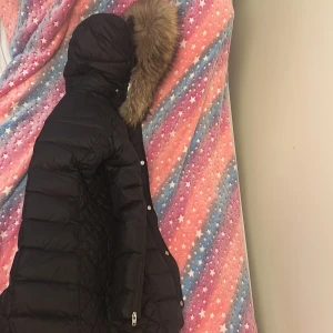 Svart dunjacka med fuskpäls från Rockandblue - Svart lång dunjacka från Rockandblue med quiltade detaljer och avtagbar huva med fuskpäls. Jackan har dragkedja, knappar och snörning i huvan. Perfekt för kalla dagar och har en snygg badge på ärmen. Vill också tillägga att den är knappt använd, den har alltså en jätte bra skick. Orginal priset är faktiskt 4000 då jag köpte den på miinto. 
