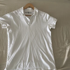 Vit pikétröja från Hampton Republic - Klassisk vit pikétröja från Hampton Republic med krage och korta ärmar. Tröjan har en stilren design med diskret logga på bröstet och knappar vid halsen. Perfekt för en clean och fräsch look under vår och sommar.