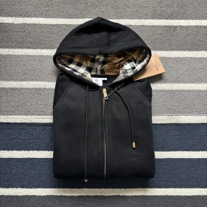 Svart Burberry hoodie med dragkedja - Svart hoodie från Burberry med klassiskt rutigt foder i huvan och guldfärgade detaljer på dragkedjan. Tröjan har långa ärmar, snörning i huvan och är tillverkad i mjuk bomull. Perfekt för dig som vill ha en stilren och lyxig look.