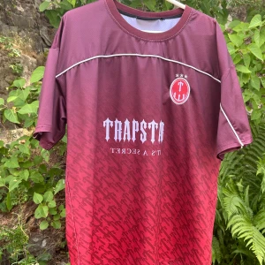 Trapstar vinröd fotbollströja M - Snygg vinröd fotbollströja från Trapstar med vit logga och text på bröstet samt nummer 22 på ryggen. Tröjan har korta ärmar, rund hals och är gjord i ett lätt, ventilerande polyestermaterial. Mönstret är diskret och ger en unik look. 