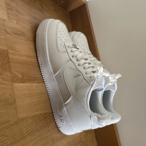 Nike Air Force 1 helvita sneakers - Klassiska Nike Air Force 1 sneakers i helvitt skinn med perforerad tå, rund tå och platt sula. Snygg och clean look med vita snören och diskret Nike-logga på hälen. Perfekta för dig som vill ha en tidlös och fräsch stil.