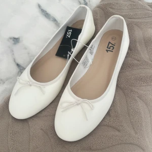 Vita ballerinaskor med rosett från 157 - Snygga vita ballerinaskor från 157 med rund tå och liten rosett framtill. Skorna har platt sula i brunt och är tillverkade i syntetmaterial. Perfekta för en clean och stilren look.