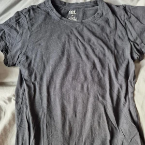 Mörkgrå t-shirt - En basic mörkgrå/svart t-shirt från Lager 157 i 100% bomull. Klassisk rund halsringning och korta ärmar, perfekt för en enkel och avslappnad stil. T-shirten är helt enfärgad utan tryck eller mönster.