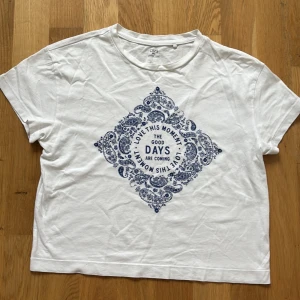 Vit t-shirt med blått tryck från OVS - Vit t-shirt från OVS med korta ärmar och rund hals. Framsidan har ett stort blått paisleyinspirerat tryck med texten 'LOVE THIS MOMENT, THE GOOD DAYS ARE COMING' i mitten. Skön passform och tillverkad i mjuk bomull. Passar för barn 12-13 år storlek 158.