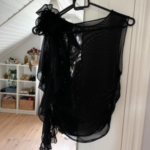 Svart transparent volangtopp med blomma - Säljer en svart, transparent topp i mesh med volangdetaljer och en stor dekorativ rosett. Perfekt att styla, använd fåtal gånger 