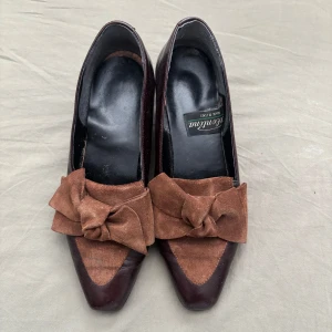 Bruna Valentinas pumps med rosett - Unika pumps från Valentina i mörkbrunt skinn med bred klack och spetsig tå. Framtill finns en stor rosett i brunt mocka som ger en cool vintagekänsla. Perfekta för dig som gillar retrostil och vill sticka ut med snygga detaljer.