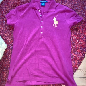 Rosa pikétröja från Ralph Lauren - Säljer en snygg rosa pikétröja från Ralph Lauren, modell The Skinny Polo. Tröjan har klassisk krage, korta ärmar och knappar framtill. Stor broderad gul logga på bröstet. Tillverkad i 100% bomull för en skön känsla. Perfekt för en sportig och stilren look.