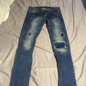 Dsquared2 slim jeans med slitningar - Blå slim fit jeans från Dsquared2 med coola slitningar och lagade hål på knäna. Byxorna har en snygg tvätt, färgstänk och en orange Dsquared2-patch bak. Tillverkade i klassisk denim med raka ben och trendig look. passar 29 i midjan