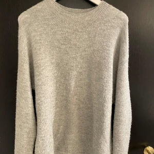 Grå stickad tröja från H&M - Säljer en stilren grå stickad tröja från H&M. Tröjan har en klassisk rund hals och långa ärmar, perfekt för kyligare dagar. Den är mjuk och bekväm, idealisk för både vardag och avslappnade tillfällen.