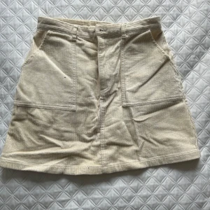 Beige manchesterkjol med fickor - Snygg beige manchesterkjol med praktiska fickor framtill. Kjolen har en klassisk A-linjeform och stängs med dragkedja. Perfekt för en stilren och avslappnad look.