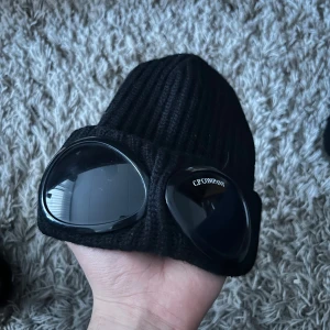 Svart mössa med goggles från C.P. Company - Säljer en svart ribbstickad mössa från C.P. Company med inbyggda goggles framtill. Ikonisk design med märkets logga på ena glaset. Köpte mössan runt 2020 och den används aldrig. Storleken är uni vilket innebär att det ska passa alla med den stretchiga materialet och att mössan kommer att förstoras i ditt storlek när den används. Nypris var 1500