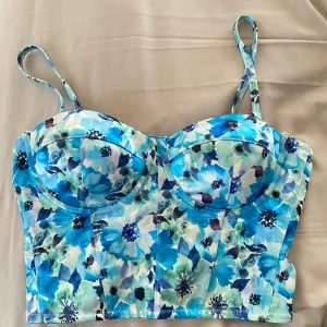 Blommig blå korsettopp - Säljer en snygg korsettopp med blått blommönster och smala axelband. Toppen har kupor och dragkedja bak. Perfekt till sommaren. Nästan aldrig använd. 💕