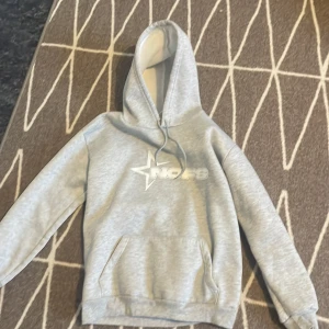 Grå hoodie med tryck - Säljer en grå hoodie med vit insida på huvan och ett tryck med stjärna och text på bröstet. Tröjan har magficka och dragsko i huvan. Perfekt för en avslappnad stil.