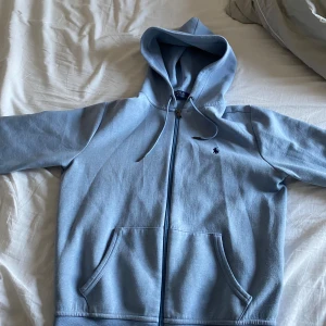 Ralph Lauren zip hoodie - Säljer en ljusblå hoodie från Polo Ralph Lauren med dragkedja och fickor framtill. Klassisk broderad logga på bröstet och justerbar huva med snören. Perfekt för en avslappnad stil. Köpt i en ralph Lauren butik skick 10/10 Inga tecken på användning, nypris ca 2300 