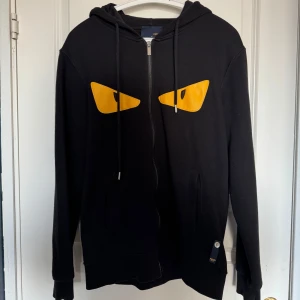 Svart hoodie med gula ögon från Fendi - Riktigt fet hoodie från Fendi med ikoniska Monster Eyes-designen i gult. Full zip-modell med huva och högkvalitativ bomull. En statement piece som kombinerar lyx med street – perfekt för dig som vill sticka ut med stil.  Ps.. liten i storlek