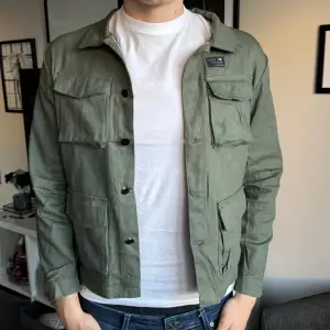 Säljer en stilren grön overshirt  med fyra stora fickor framtill och knäppning med svarta knappar. Jackan har klassisk krage och är perfekt att bära över en t-shirt för en avslappnad look.