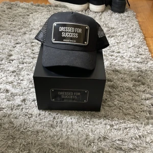 Svart keps från John Hatter & Co - Snygg svart keps med texten 'Dressed for Success' på framsidan. Kepsen har ett nätmaterial på baksidan för bättre ventilation och justerbar passform. Perfekt för sommaren! Aldrig använd!