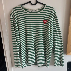  Comme des Garçons långarmad - Säljer en stilren randig tröja från Comme des Garçons i grönt och vitt. Tröjan har ett ikoniskt rött hjärta på bröstet och långa ärmar. Perfekt för en avslappnad och trendig look.