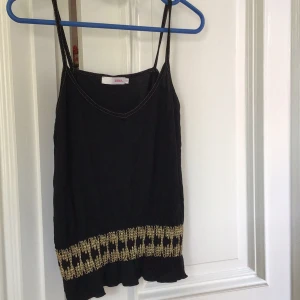 Svart linnetopp från Zoul - Snygg svart linnetopp från Zoul med smala axelband och dekorativa guldfärgade broderier längs nederkanten. Perfekt för en stilren look.