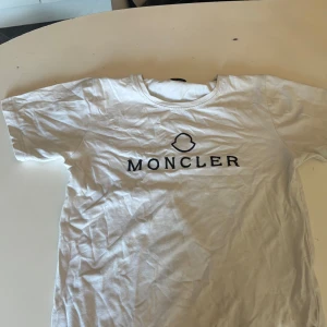 Vit t-shirt från Moncler - Säljer en klassisk vit t-shirt från Moncler med deras ikoniska logga tryckt i svart på bröstet. T-shirten har en rund halsringning och korta ärmar. Perfekt för en stilren och avslappnad look.