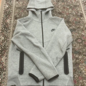 Grå hoodie från Nike - Säljer en stilren grå hoodie från Nike med dragkedja och huva. Tröjan har långa ärmar och svarta detaljer vid fickorna. Perfekt för en avslappnad look.
