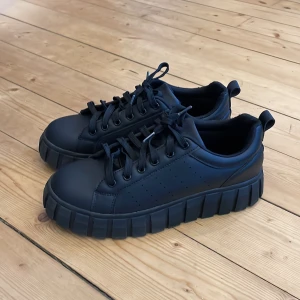 Svarta sneakers med grov sula - Snygga svarta sneakers med en rejäl och grov sula. Skorna har snörning och en rund tå, vilket ger en cool och modern look. Perfekta för dig som vill ha en stilren men ändå iögonfallande sko.