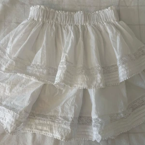 Vit volangkjol med spetsdetaljer - Supersöt vit kjol med dubbla volanger och fina spetsdetaljer. Kjolen har resår i midjan och är tillverkad i ett lätt och luftigt material, perfekt för varma dagar.