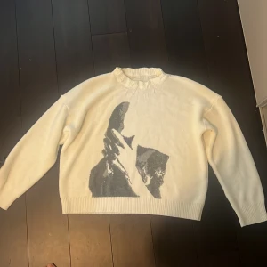 Vit stickad tröja med tryck - Säljer en vit stickad tröja med ett unikt tryck i grått på framsidan. Tröjan har en ribbad krage och långa ärmar, perfekt för kyligare dagar. Den är både stilren och bekväm.