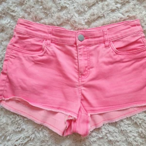 Rosa jeansshorts med rå kant - Säljer ett par ljusrosa jeansshorts med råa kanter och klassisk femficksdesign. Shortsen har knapp och dragkedja framtill samt bälteshällor. Perfekta för varma dagar och enkel att matcha med olika toppar.