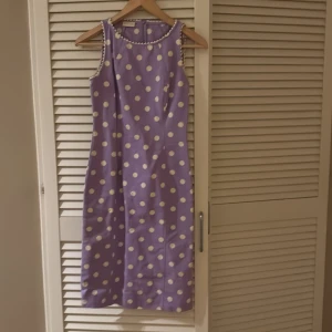 Lila prickig klänning från Valentino - Superfin lila klänning från Valentino med vita prickar och dekorativ kant i vitt runt hals och ärmhål. Klänningen är ärmlös och har en rak, klassisk passform som slutar vid knäna. Perfekt för dig som vill sticka ut med en retroinspirerad look.