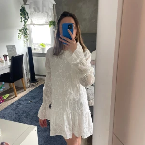 Studentklänning  - Supersöt vit klänning med blommigt broderi, volangkant nertill och långa ärmar. Klänningen har en öppen rygg med knytband i nacken och är tillverkad i ett lätt, luftigt material. Perfekt för sommarens alla tillfällen!