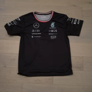 Svart Mercedes AMG Petronas t-shirt - Sportig svart t-shirt från Mercedes AMG Petronas med tryckta loggor från sponsorer på både fram- och baksida. T-shirten har rund hals med röd och vit kant samt korta ärmar. Perfekt för motorsportfans.