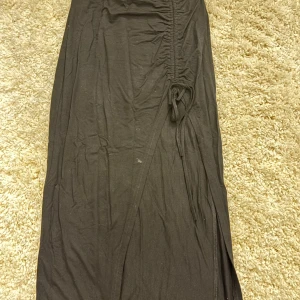Svart långkjol med dragsko - Svart långkjol med snygg knytning i sidan och en slits som ger extra stilpoäng. Kjolen har en rak passform och är gjord i ett mjukt bomullsmaterial. Den har en liten fläck som jag inte testat att tvätta bort, den syns ej så mycket. 