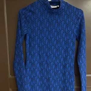 Blå långärmad klänning med mönster  - Säljer en blå långärmad klänning från junkyard med svart grafiskt mönster. Klänningen har hög hals och asymmetrisk nederkant. Perfekt för dig som gillar unika detaljer och vill sticka ut.