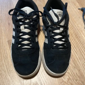 Svarta Adidas sneakers med vita ränder - Säljer ett par klassiska svarta Adidas sneakers med vita ränder på sidorna och vit sula. Skorna har snörning och rund tå. Perfekta för vardagsbruk och har en tidlös design.