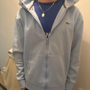 Ljusblå kofta från Lacoste - Säljer en ljusblå kofta från Lacoste med dragkedja framtill och två fickor. Perfekt för en avslappnad stil.