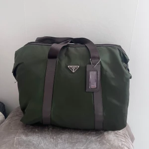 Mörkgrön weekendbag från Prada - Säljer en snygg mörkgrön weekendbag från Prada med bruna läder detaljer och dubbla handtag. Väskan har klassisk trekantig Prada-logga framtill och är tillverkad i slitstark nylon med läderdetaljer. Perfekt för resor eller gymmet. Passar perfekt som handbagage, med korrekta mått, 35x45x15