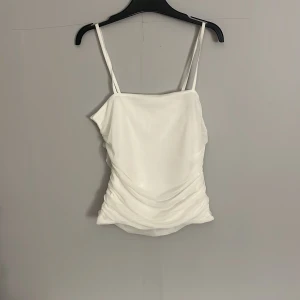 Vit omlott linnetopp från Gina Tricot - Säljer en vit linnetopp från Gina Tricot med smala axelband och snygga draperade detaljer på sidorna. Toppen har en omlott-effekt och är perfekt för sommaren. Materialet känns lätt och luftigt.