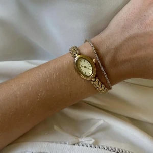 Bovou klocka  - Elegant guldfärgad klocka från Bovou med oval urtavla i vitt och länkarmband i metall. Klockan har minimalistisk design med tunna visare och indexmarkeringar. Perfekt accessoar för en stilren look. Tillkommer avtagna länkar och box samt verktyg för att ändra länkarna. Nypris är 1400kr