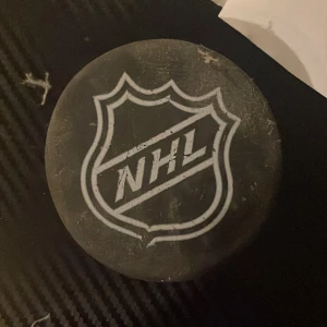 NHL hockeypuck - Säljer en klassisk svart hockeypuck med NHL-loggan tryckt i vitt på ena sidan. Pucken är rund och tillverkad i hårt gummi, perfekt för hockeyträning eller som samlarobjekt för NHL-fans.