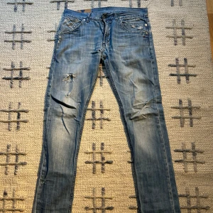 Blå jeans med slitningar - Säljer ett par blå jeans med coola slitningar och en klassisk femficksdesign. Jeansen har en dragkedja och knapp framtill. Perfekta för en avslappnad stil. Skickt 4/10 