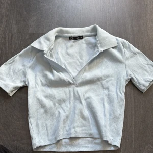Ljusblå ribbad pikétröja från Zara - Säljer en ljusblå pikétröja från Zara. Tröjan är ribbad med korta ärmar och en krage. Använd en gång så nästan ny, den sitter tajt i både ärm och runt kroppen samt den har en V ringning 